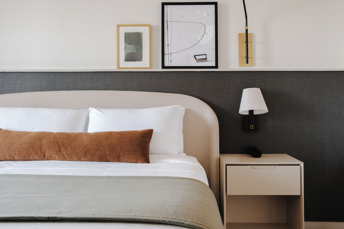 A neatly made bed with white and brown pillows, a bedside table with a lamp, and two framed pictures on the wall.