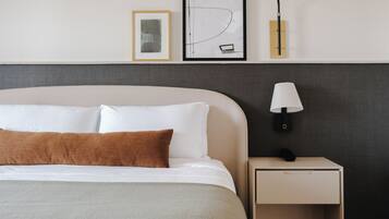 Una cama bien hecha con almohadas blancas y marrones, una mesita de noche con lámpara, y dos cuadros enmarcados en la pared.