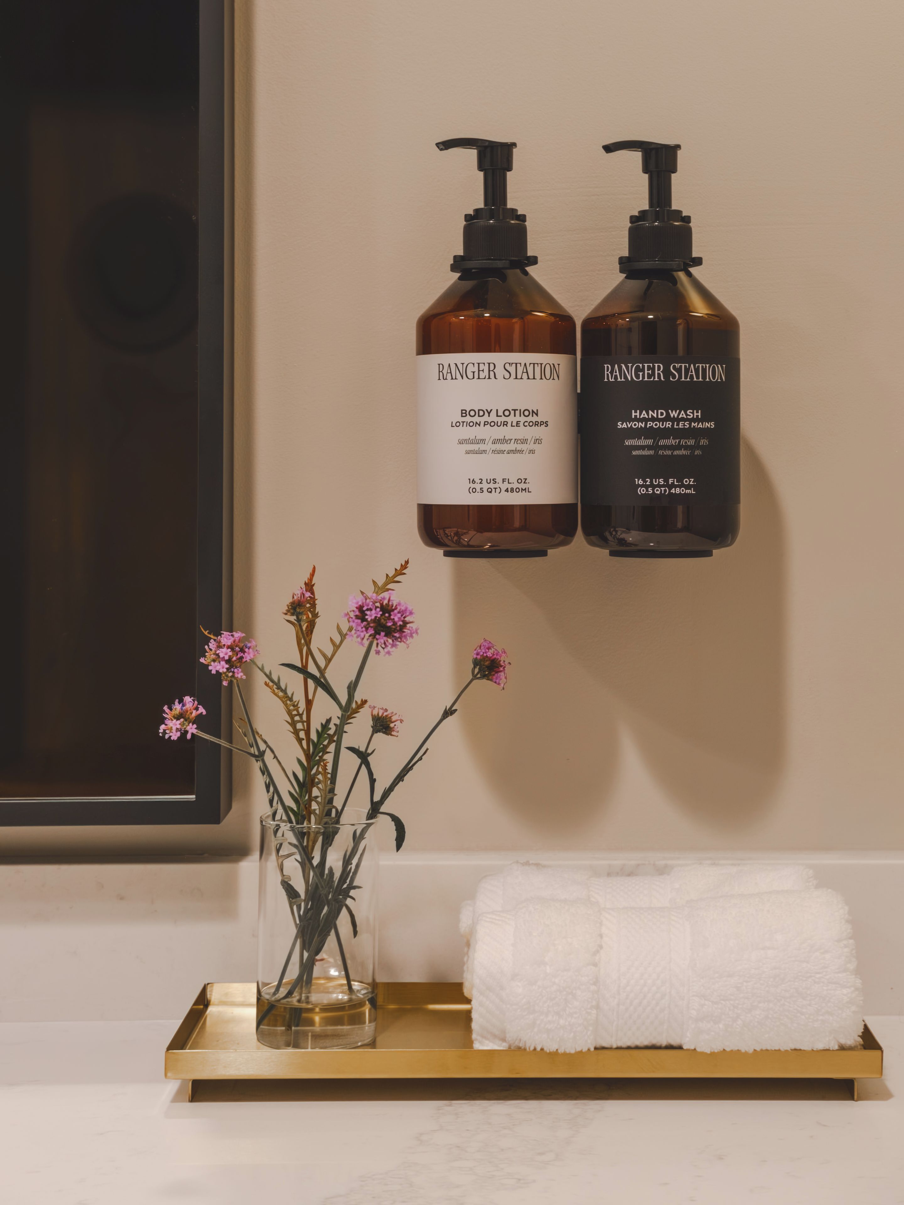 Et badeværelse med to flasker Ranger Station body lotion og håndvask på en hylde, en vase med blomster og et hvidt håndklæde.