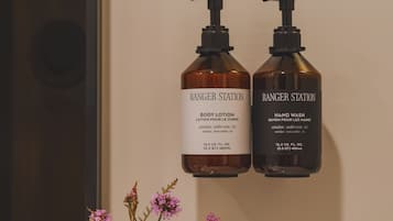 Et badeværelse med to flasker Ranger Station body lotion og håndvask på en hylde, en vase med blomster og et hvidt håndklæde.