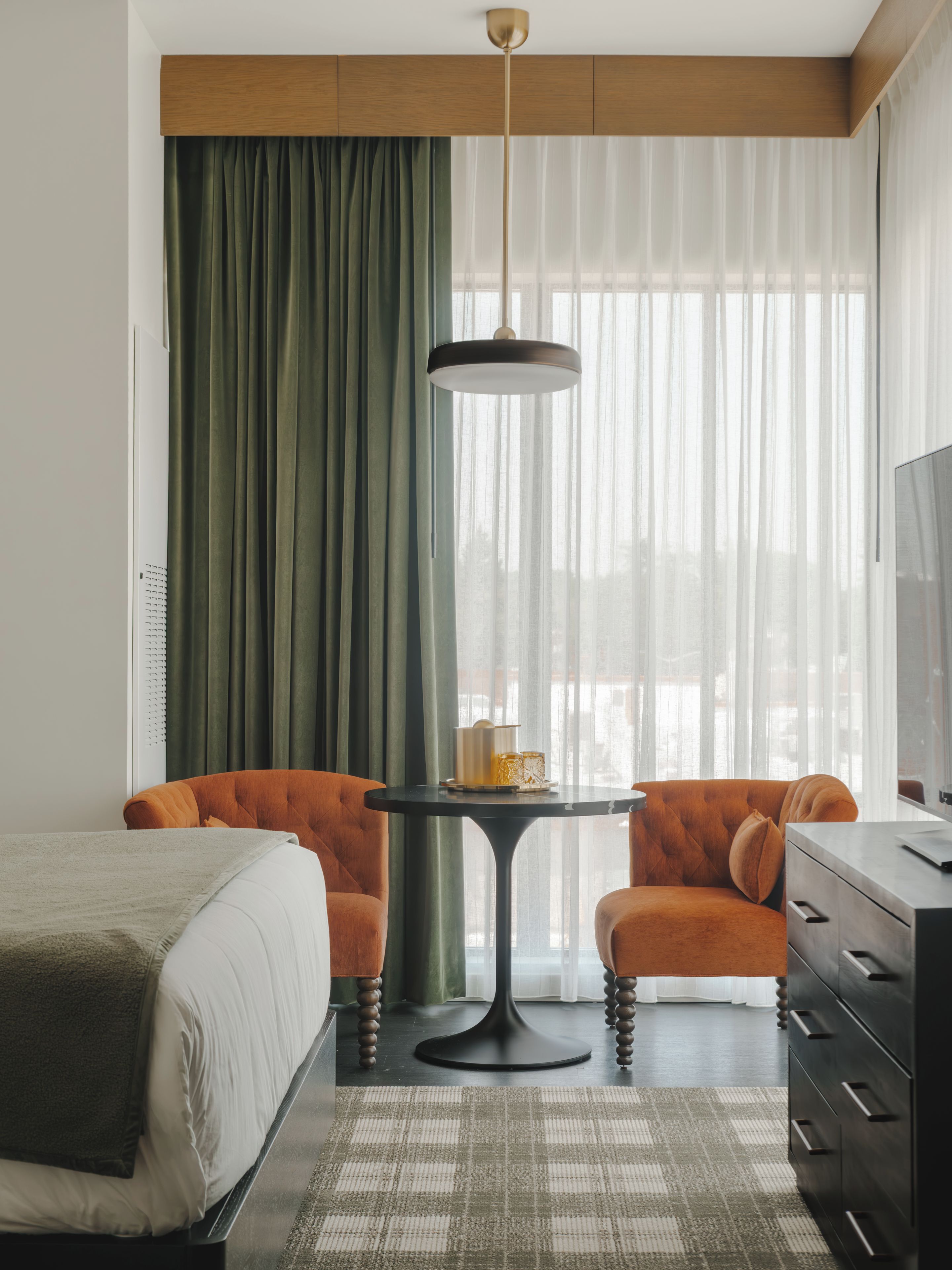 Une chambre d’hôtel comprenant un lit, deux fauteuils, une petite table, une commode et une grande fenêtre avec des rideaux légers.