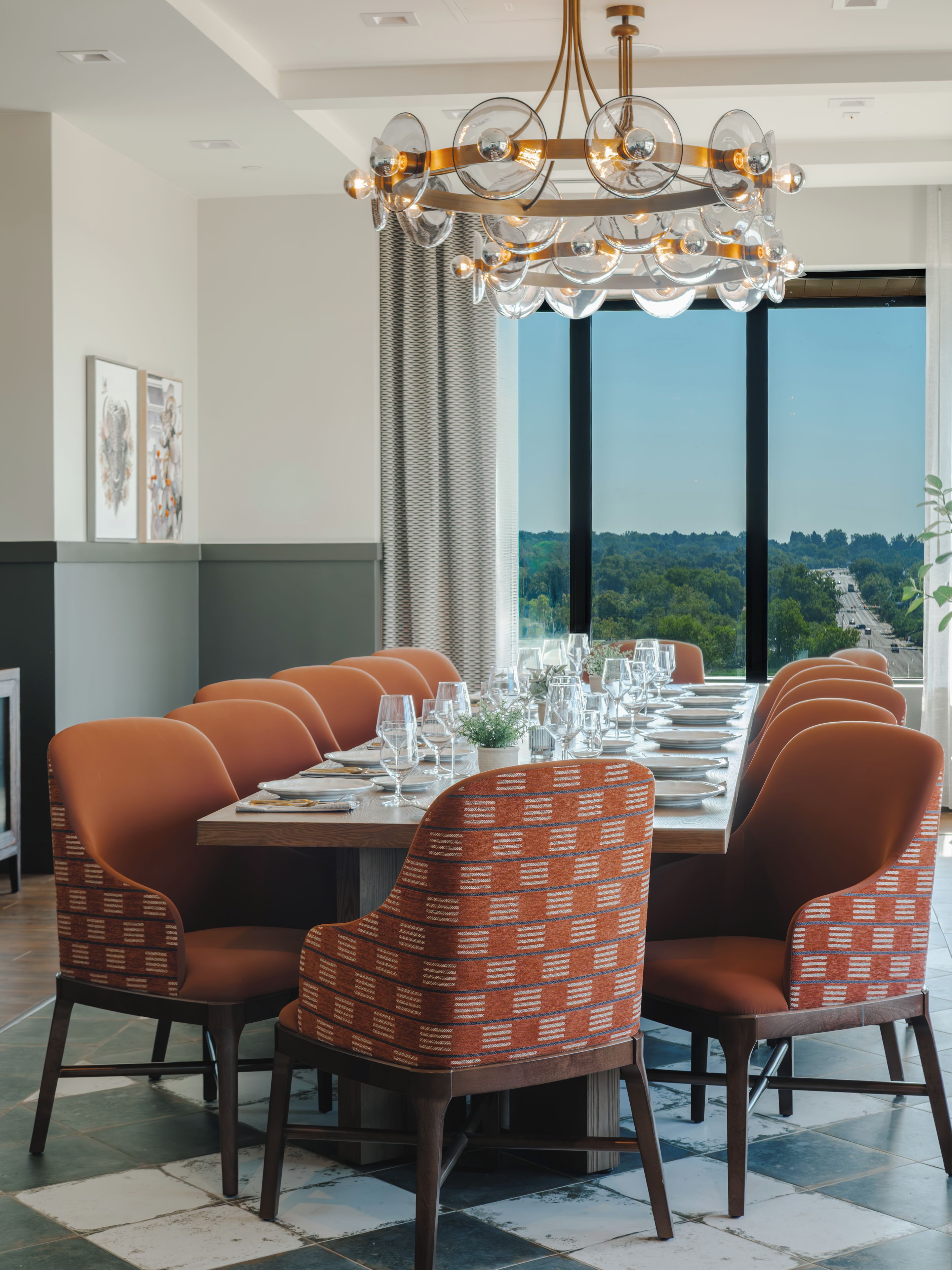 Un comedor con una mesa larga dispuesta para seis personas, rodeada de sillas estampadas, una moderna lámpara de araña y vistas a un paisaje verde a través de amplios ventanales.
