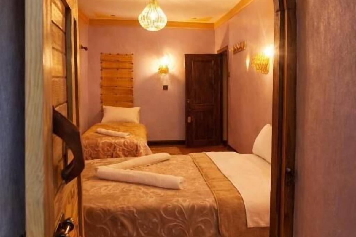Una habitación con una cama, una puerta de madera, una araña y una lámpara de pared.