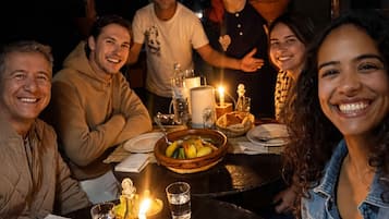 Eine Gruppe von Menschen speist an einem Tisch im Freien, mit Kerzen, Tellern und einer Obstschale.