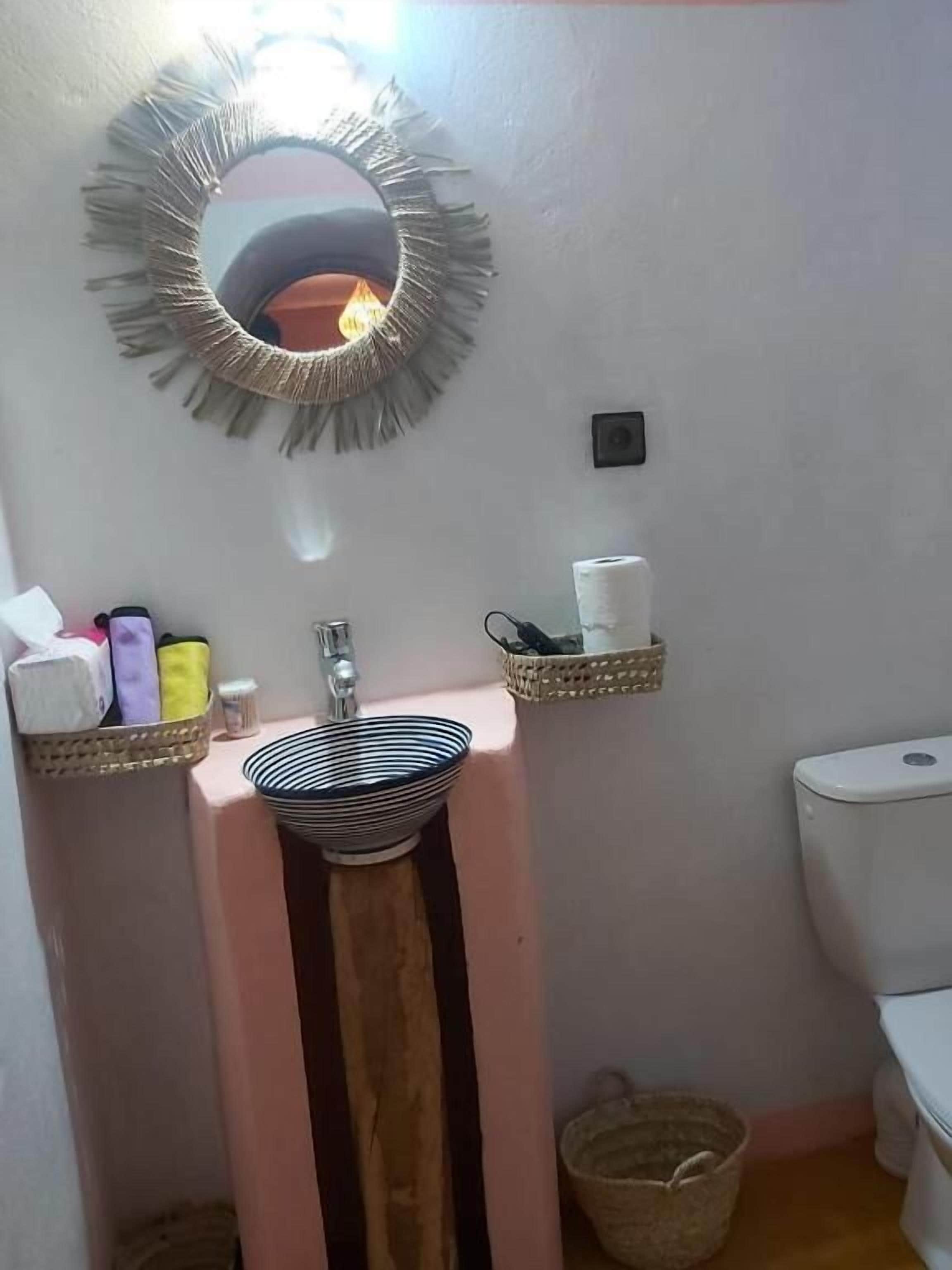 Un baño con un lavabo rosa, un espejo con diseño de flecos y dos repisas con artículos de tocador.