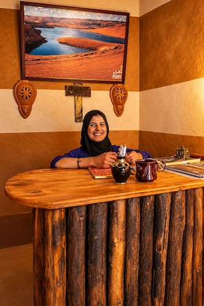 Un mostrador de recepción de madera con una persona sentada detrás, un cuadro de paisaje enmarcado encima y una cruz en la pared.