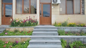 Un bâtiment doté de deux portes en bois, d’une fenêtre avec un rideau et d’un petit jardin où poussent différentes sortes de fleurs.