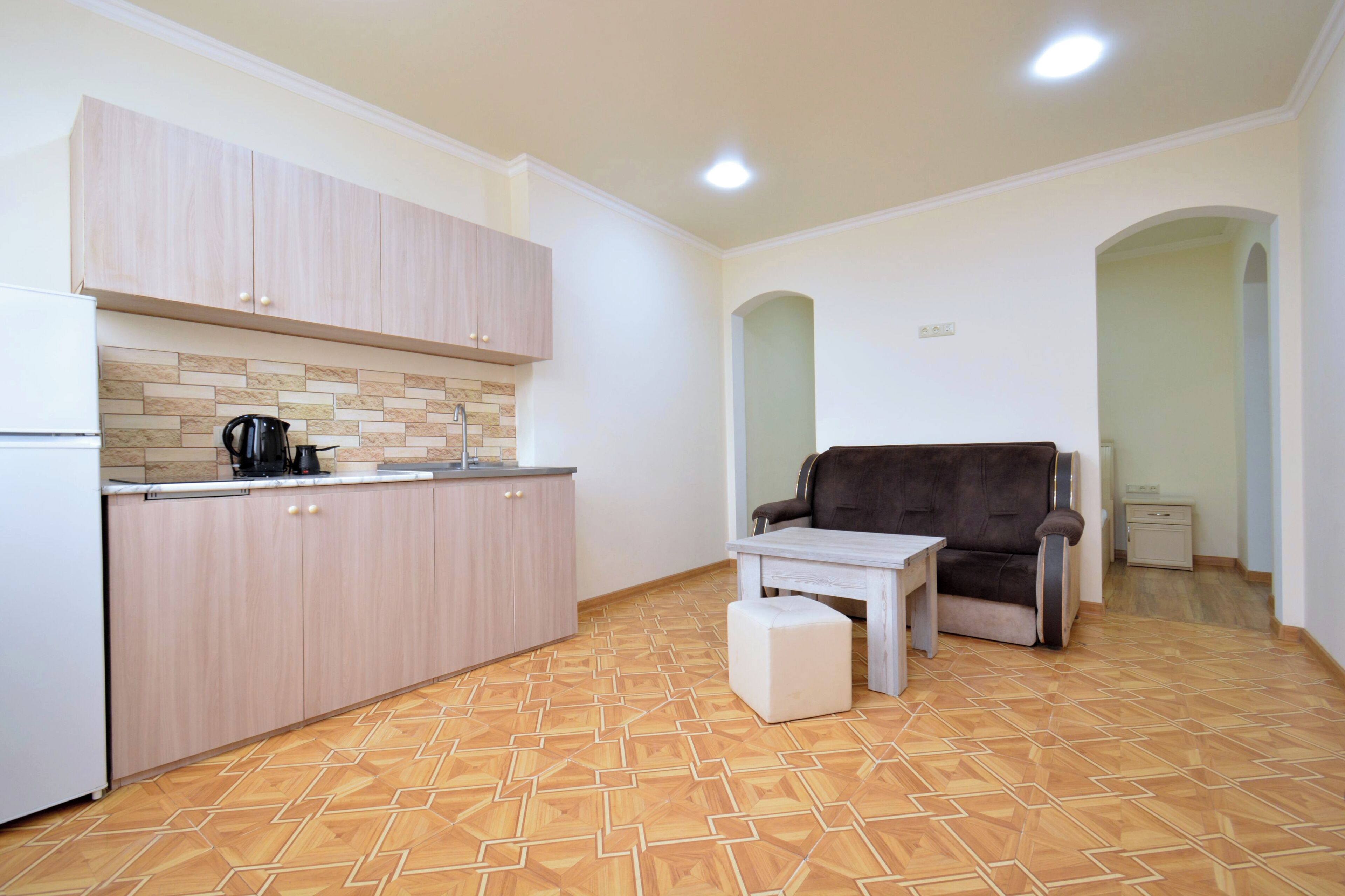 A kitchen with light wood cabinets, a tiled backsplash, a coffee maker, and a sofa in a living area.