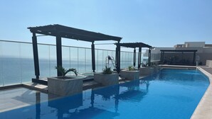 Uma piscina infinita no terraço, com guarda-corpo de vidro, plantas em vasos e vista para o oceano.
