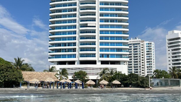 Um hotel com vários andares e varandas, situado perto de uma praia com guarda-sóis e palmeiras.