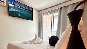 Ein Hotelzimmer mit Bett, einem an der Wand befestigten Fernseher, einem Fenster mit Vorhängen und einer schwarzen Lampe auf dem Nachttisch.