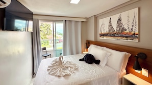 Ein Hotelzimmer mit einem Bett, einem Gemälde, einem Nachttisch, einer Lampe, einem Sessel und Blick ins Freie.