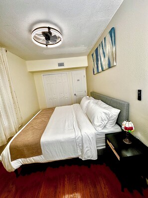 Une chambre d’hôtel avec un lit, une table de chevet, une lampe, un ventilateur de plafond et un tableau au mur.