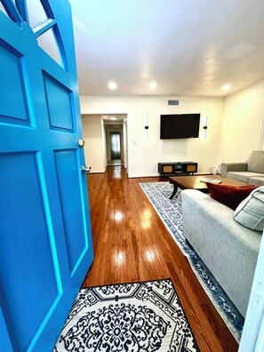 Ein Wohnzimmer mit blauer Tür, Holzdielenboden, einem grauen Sofa, einem Couchtisch, einem an der Wand montierten Flachbildfernseher und einem gemusterten Teppich.