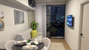 Un salon moderne avec un canapé, une table à manger dressée avec des assiettes et des serviettes, un téléviseur fixé au mur et une vue sur la ville à travers une porte vitrée.