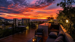 Une terrasse sur le toit, aménagée avec du mobilier d’extérieur, un espace bar et une vue panoramique sur la ville au coucher du soleil.