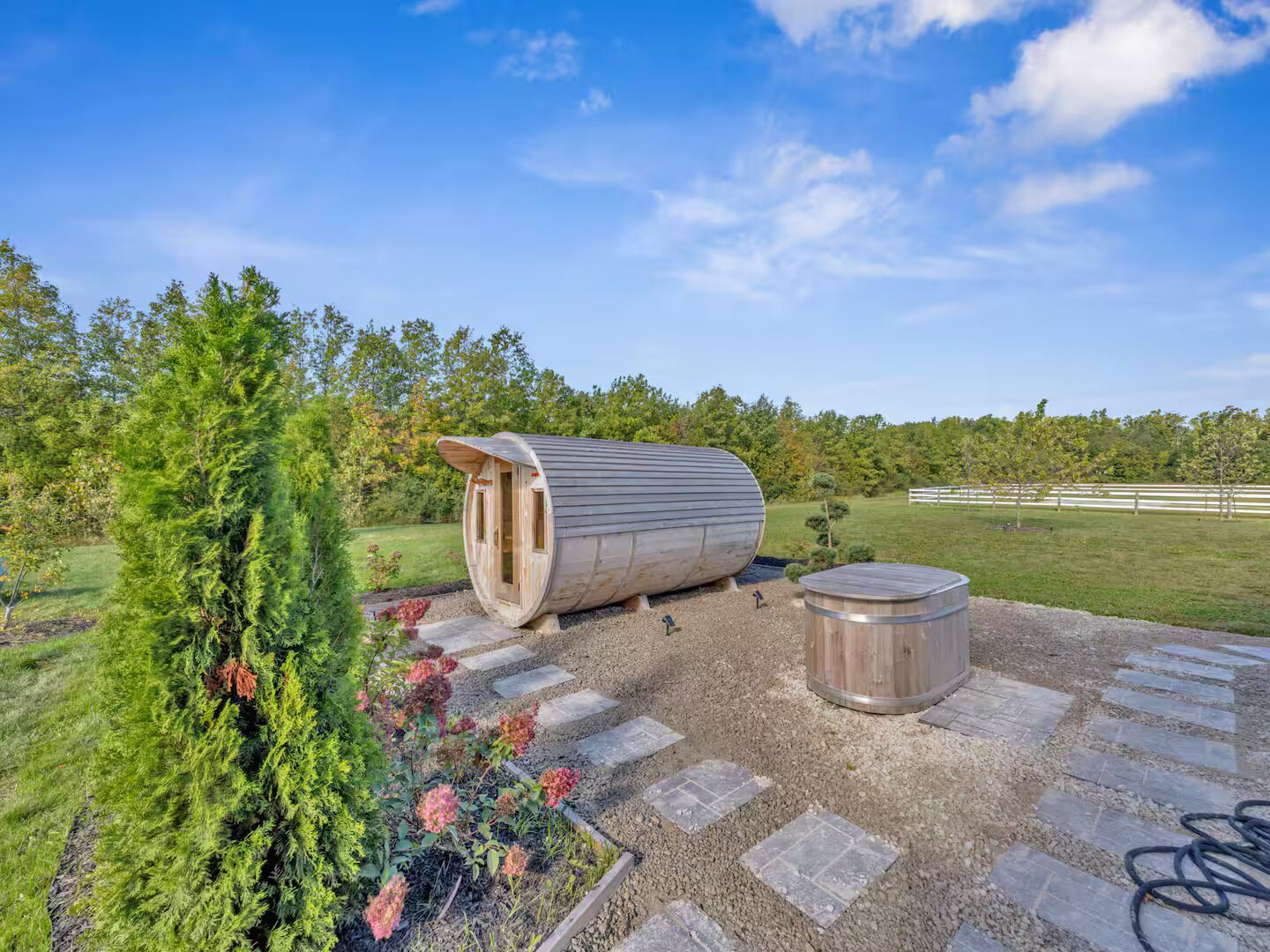 A cylindrical wooden structure with a door, a stone bench, and a garden with trees and flowers.