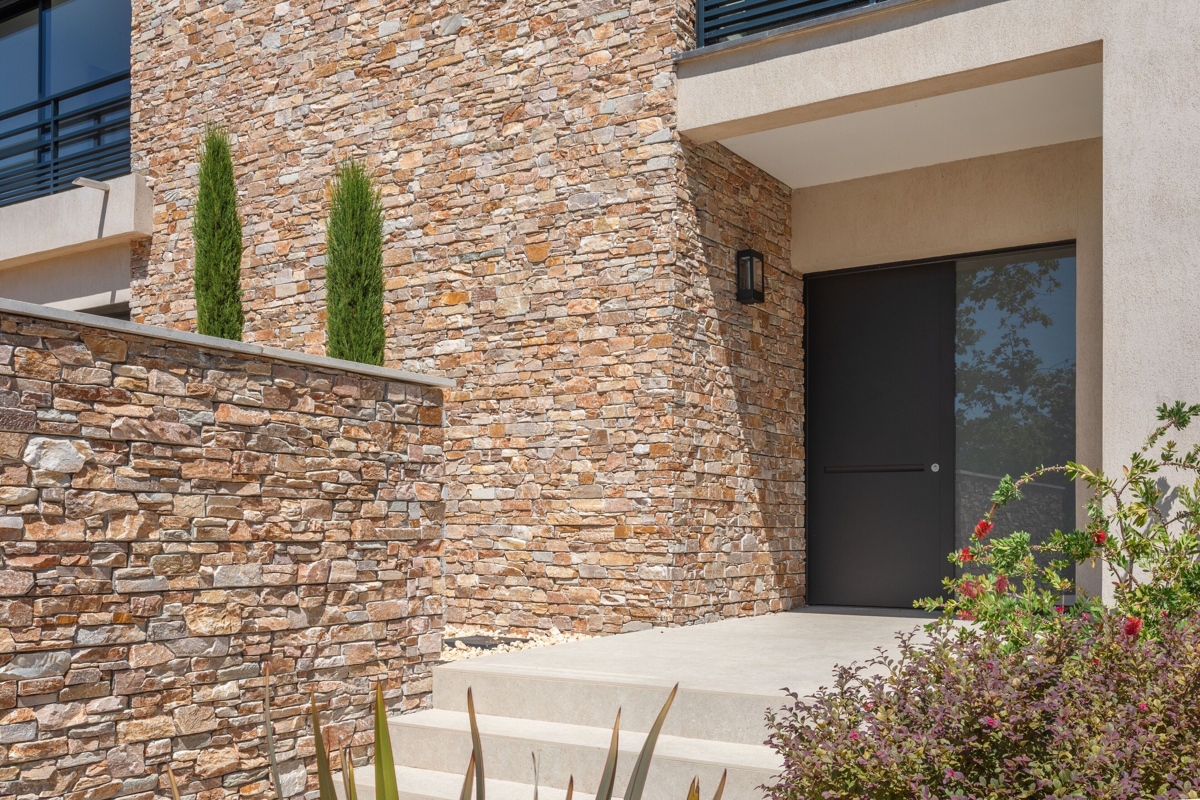 A modern building with a stone facade, a dark door, and a small garden with plants and flowers.