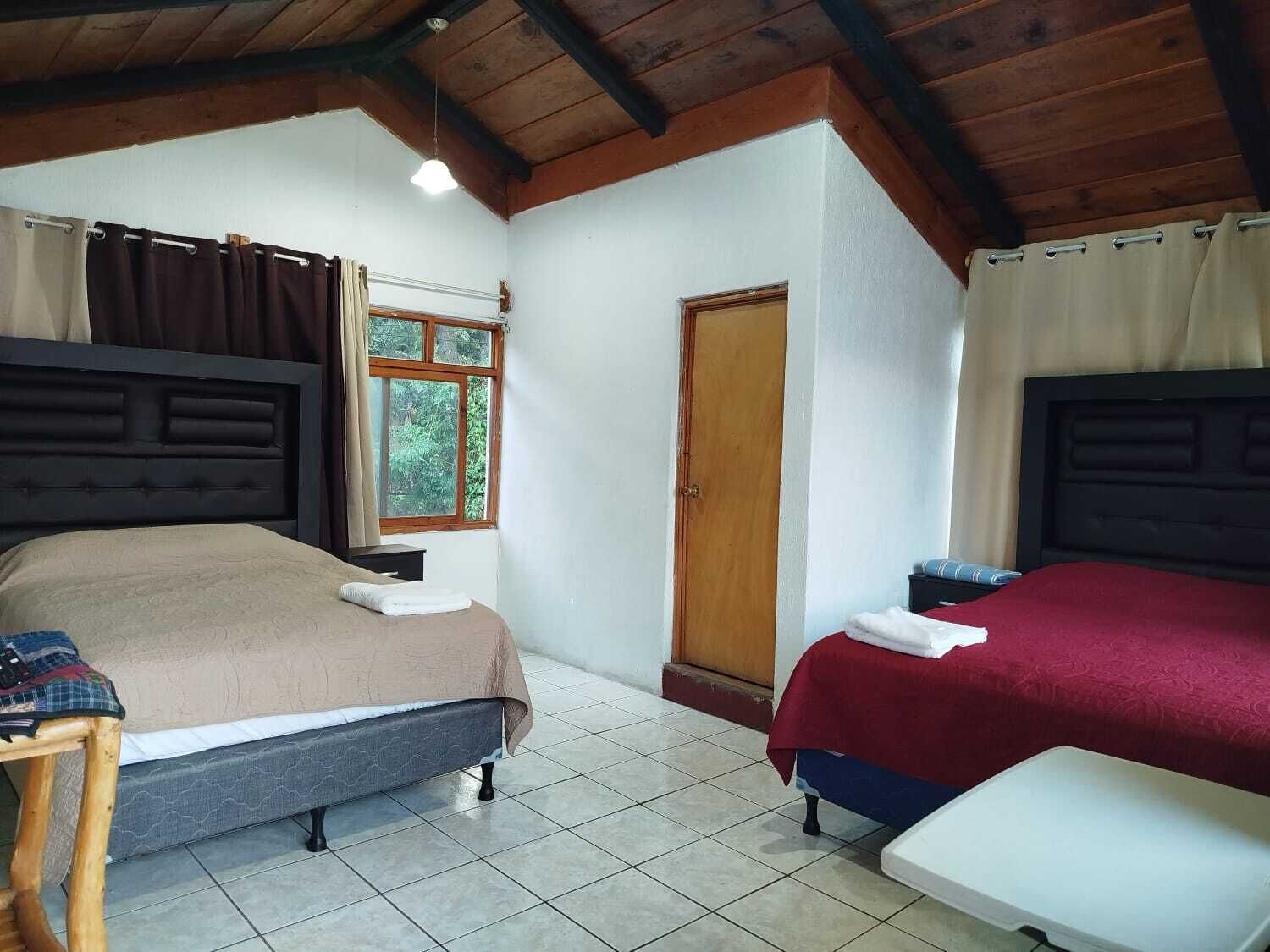 A room with two beds, a wooden ceiling, a window with curtains, and a door.