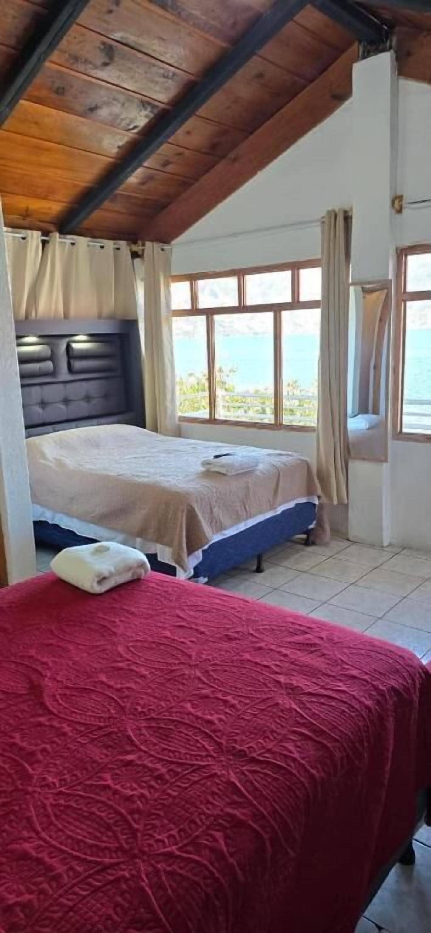 Una habitación con dos camas, una con cubrecama rojo y la otra con cubrecama color beige. El techo es de madera y se ve el mar a través de ventanas amplias.