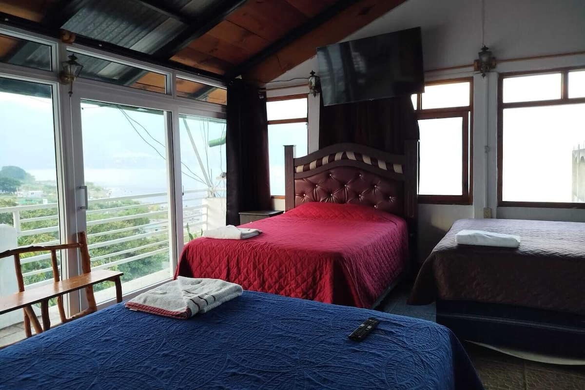 Un dormitorio con un ventanal grande, una cama con una colcha roja y otra cama con una colcha azul.
