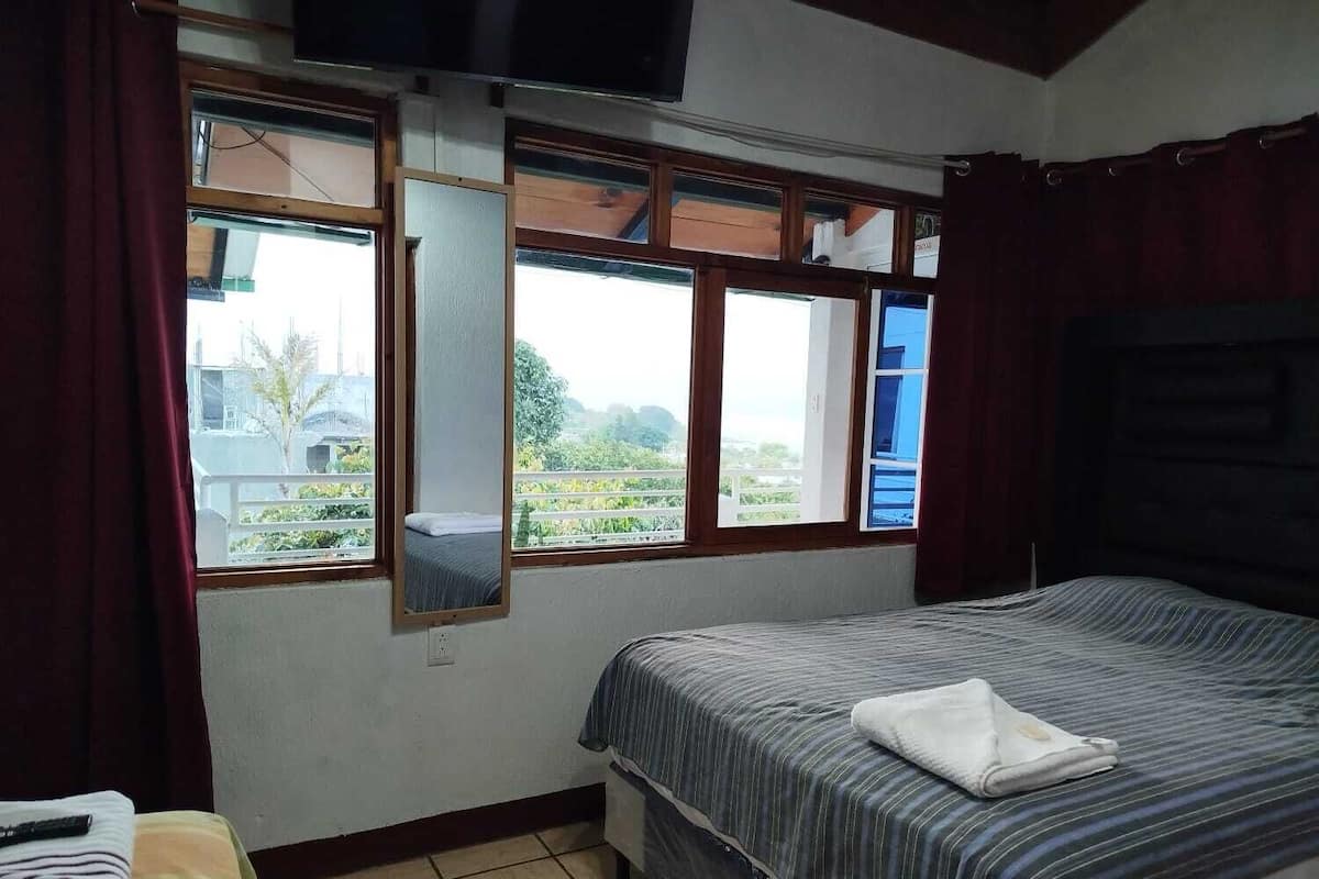 Un dormitorio con una cama, un televisor montado en la pared y una ventana con vistas al verdor exterior.