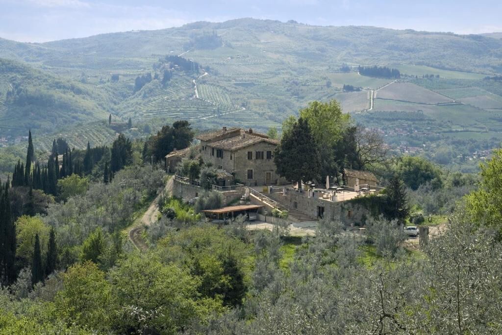 A historic stone building with a courtyard, surrounded by olive groves and hills.