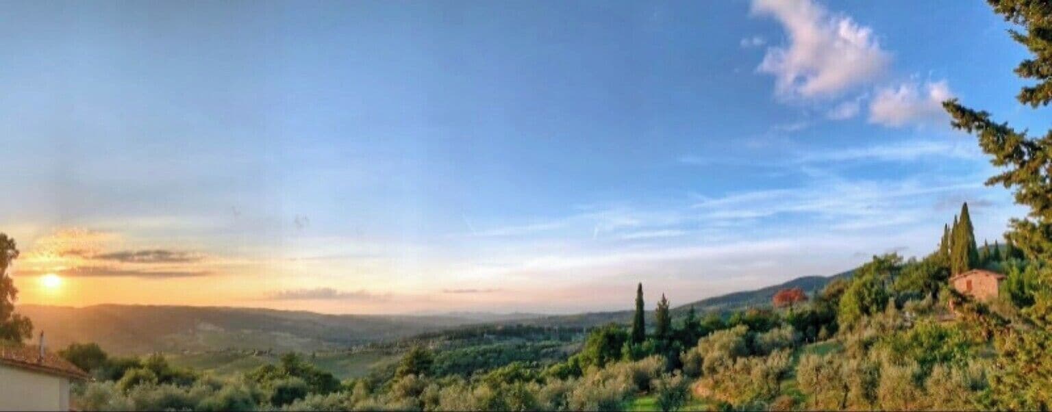 Vista panorámica de un paisaje montañoso con atardecer, árboles y un edificio distante.