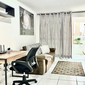 Ein modernes Wohnzimmer mit einer Couch, einem Schreibtisch mit Computer, einem Sessel und einem großen Fenster mit Vorhängen.