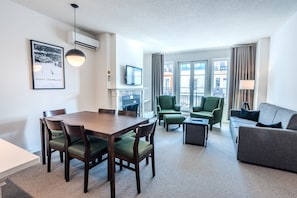 Ein Hotelzimmer mit Esstisch, grünen Polsterstühlen, einem grauen Sofa, einem Flachbildfernseher und einem Kamin.