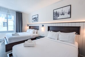 A hotel room with two beds, a large window with curtains, a framed picture on the wall, and a headboard with a light fixture.