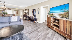 Een woonkamer met een grijze bank, een bijzettafeltje, een houten tv-meubel en een televisie waarop een strand te zien is.