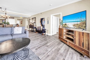 A living room with a grey sofa, a coffee table, a wooden entertainment unit, and a TV displaying a beach scene. - Tidewater 2405 (Panama City Beach)