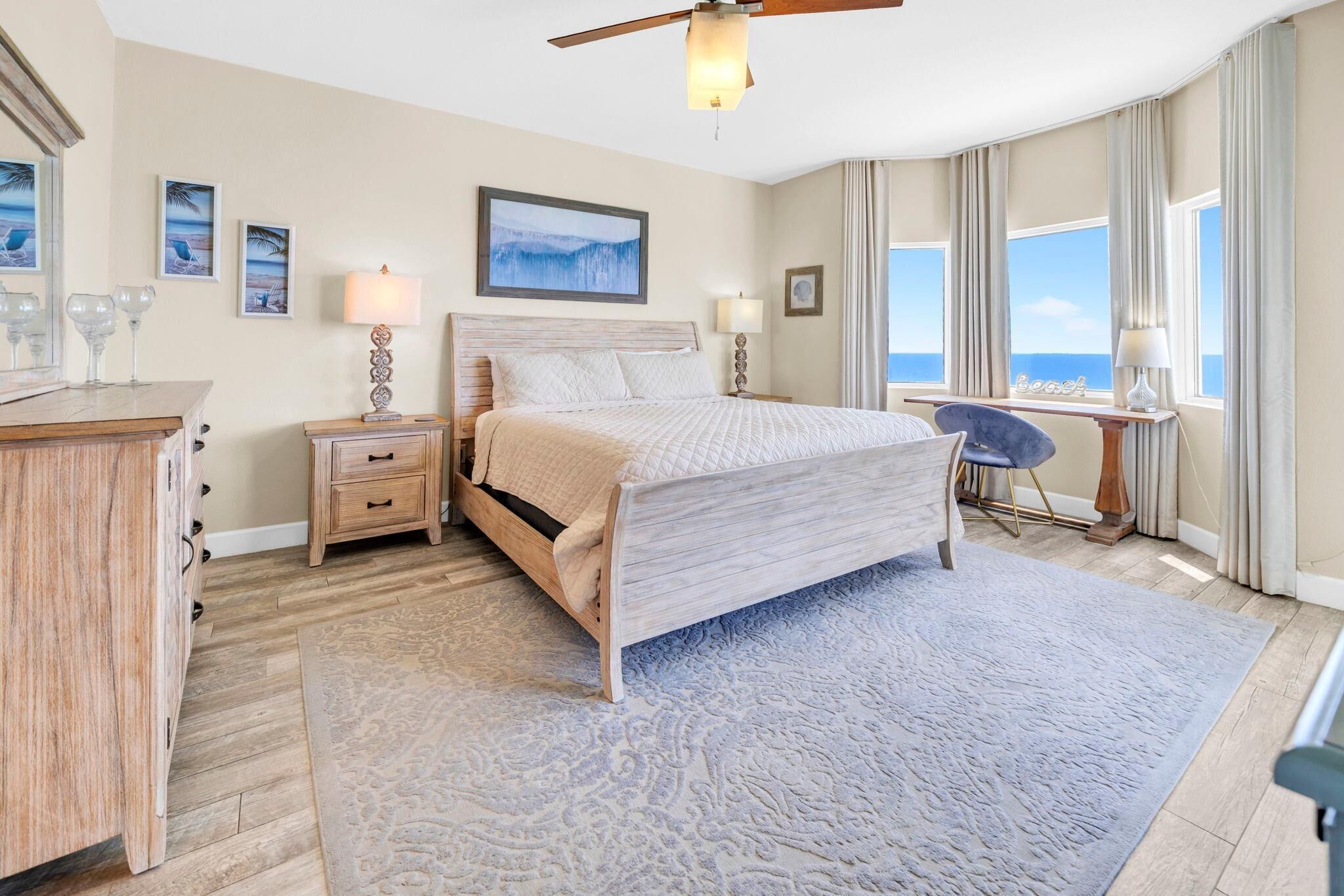 A bedroom with a bed, nightstands, a desk, and a chair. There are framed pictures on the wall, a ceiling fan, and a view of the ocean.