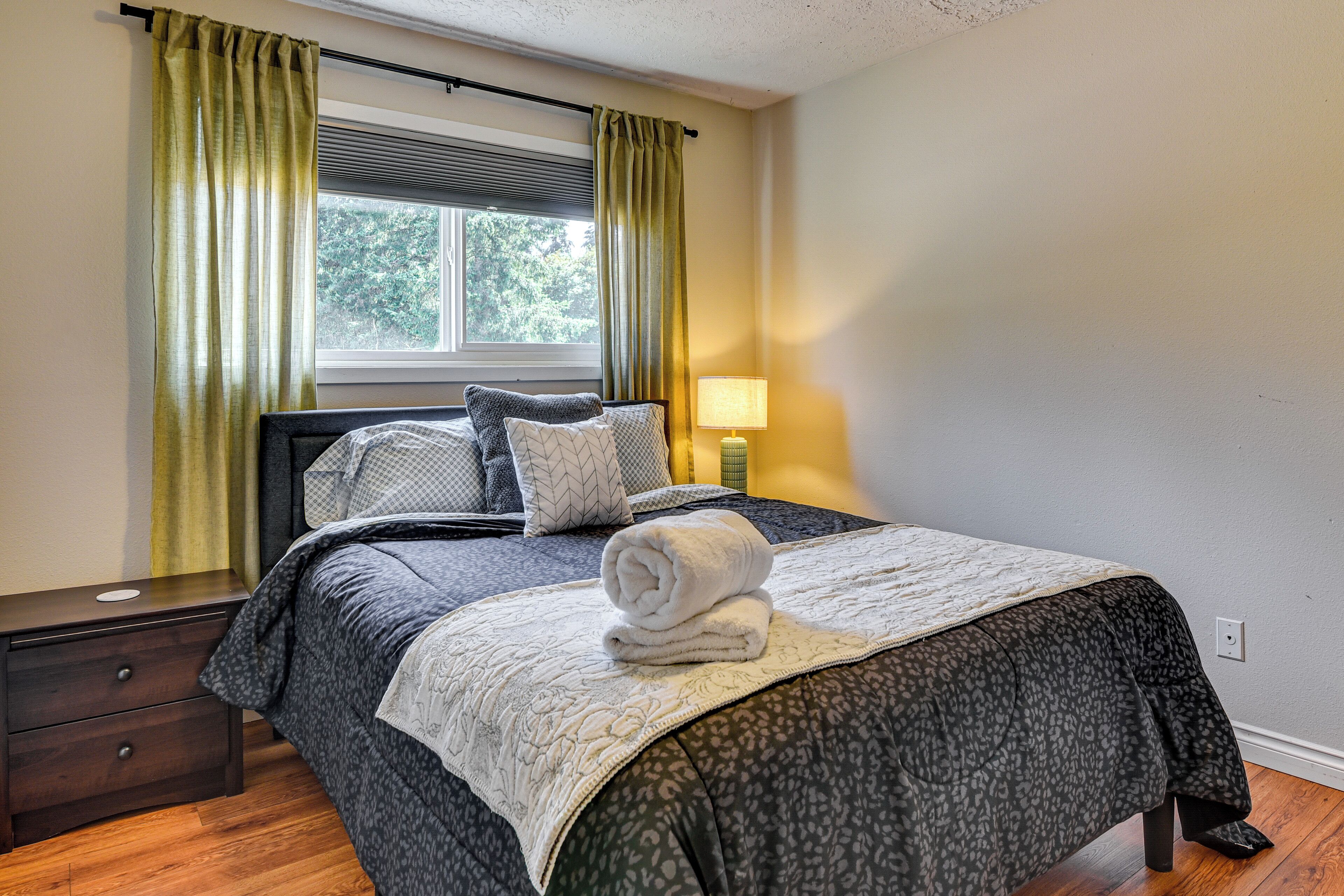 A neatly made bed with a rolled towel, a bedside lamp, and a window with curtains.