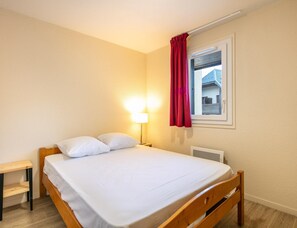 A single bed with white bedding, a bedside table, a lamp, a window with a red curtain, and a view of a house outside. - Spacious Group Accommodation in the Valley (Saint-Colomban-des-Villards)