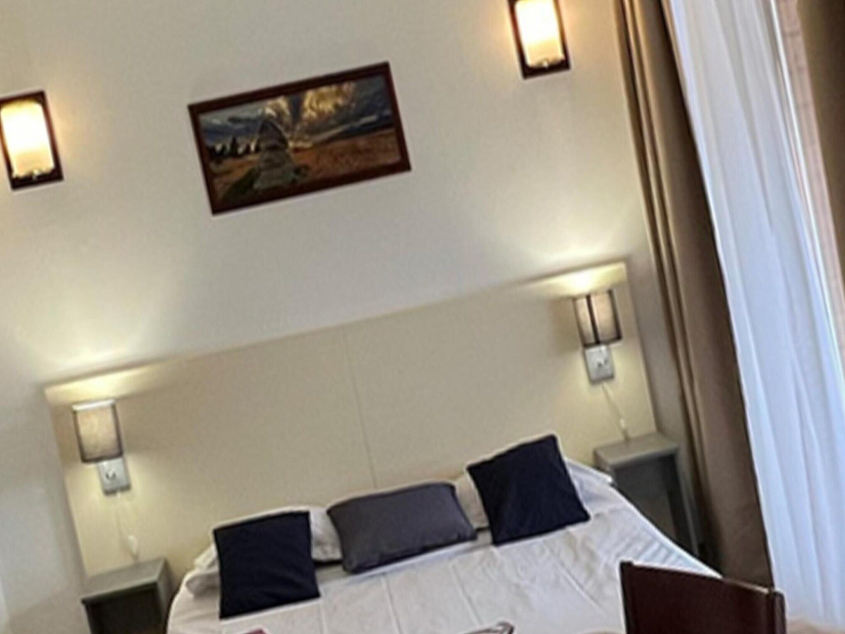 A hotel room with a bed, two bedside tables, a painting on the wall, and two wall-mounted lamps.