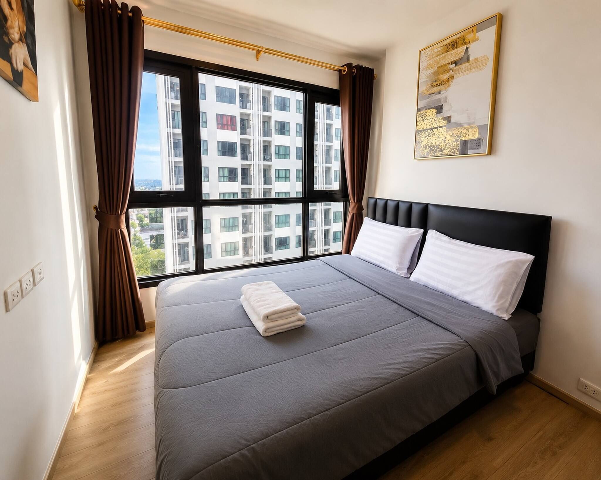 A bedroom with a large window, a bed with grey bedding, a black headboard, and a painting on the wall.