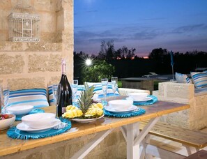 An outdoor dining setup with a stone wall, a table set with plates, glasses, and a bottle, and a birdcage on the wall. - Prestige Pool Trullo (Racale)