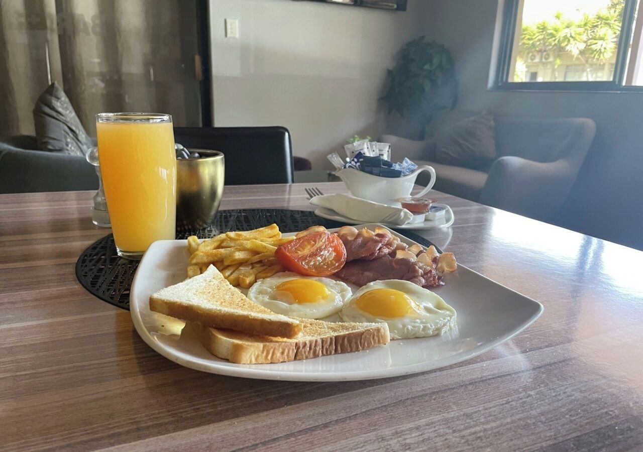 Une assiette de petit-déjeuner avec des œufs, du bacon, des tomates et des toasts, un verre de jus d’orange et une tasse de café, le tout posé sur une table.