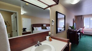 A hotel room with a large mirror, a sink, a bed with white linens, a desk with a chair, and a window with curtains.