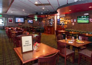A restaurant with multiple seating areas, including booths and tables. There are televisions mounted on the walls, a bar area, and a sign indicating free daily breakfast. - Queen Room w/ Free Breakfast & Airport Shuttle (Medford)