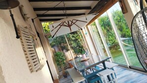 Een overdekt terras met een tafel en stoelen, een hangklok en uitzicht op de tuin.