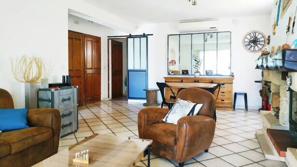 Een woonkamer met een bruine leren fauteuil, een houten salontafel, een eethoek met een houten tafel en stoelen, en een keuken met een fornuis en kasten.