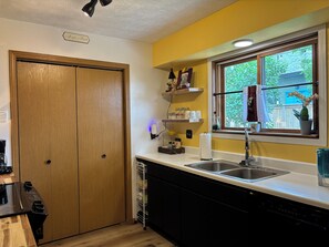 A kitchen with wooden cabinets, a dishwasher, a window with a view of greenery, and a shelf with kitchen items. - Cute & comfy, close to Gallatin County Fairgrounds & Gallatin Valley BMX Park (Bozeman)