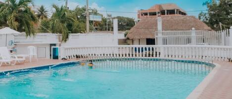 Una piscina con una cerca blanca, palmeras y un edificio con techo de paja al fondo.
