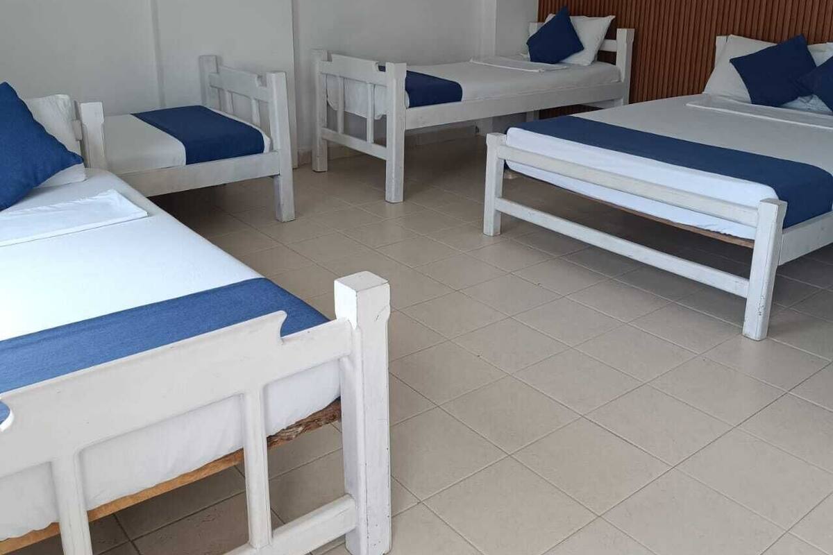 Habitación de hotel con cuatro camas blancas, cada una con ropa de cama azul, cabeceros de madera y un equipo de aire acondicionado en la pared.