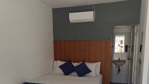 A hotel room with a bed, a wooden headboard, a white air conditioning unit, a bathroom with a sink, and a door leading to another room.