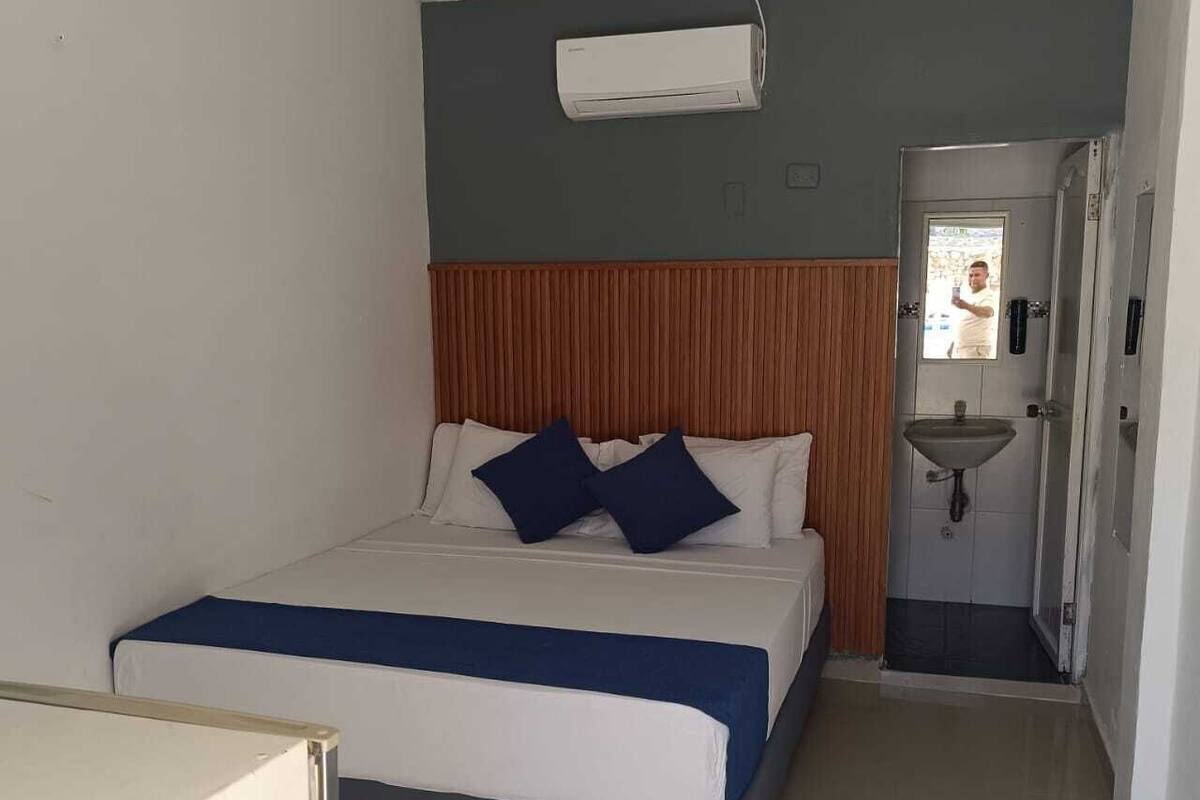 Habitación de hotel con una cama, cabecero de madera, unidad de aire acondicionado blanca, baño con lavamanos y una puerta que da a otra habitación.