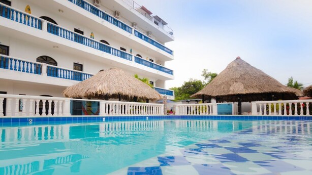A swimming pool with a checkered pattern, a white building with blue balconies, and thatched-roof structures.